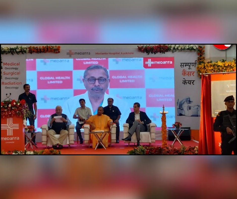 A giant screen behind them displaying a senior healthcare leader speaking virtually